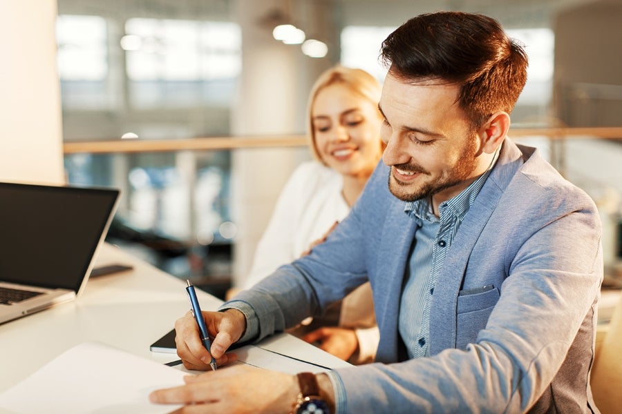 Smiling man in blue blazer signs document while blonde woman watches
