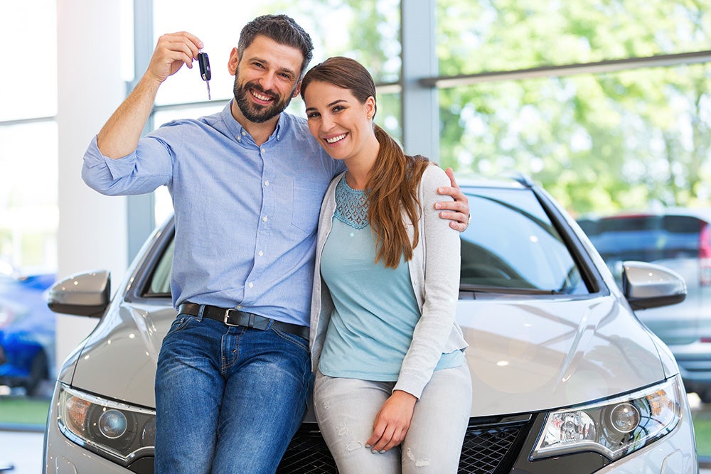 Happy couple smiling and holding keys while leaning on their new car